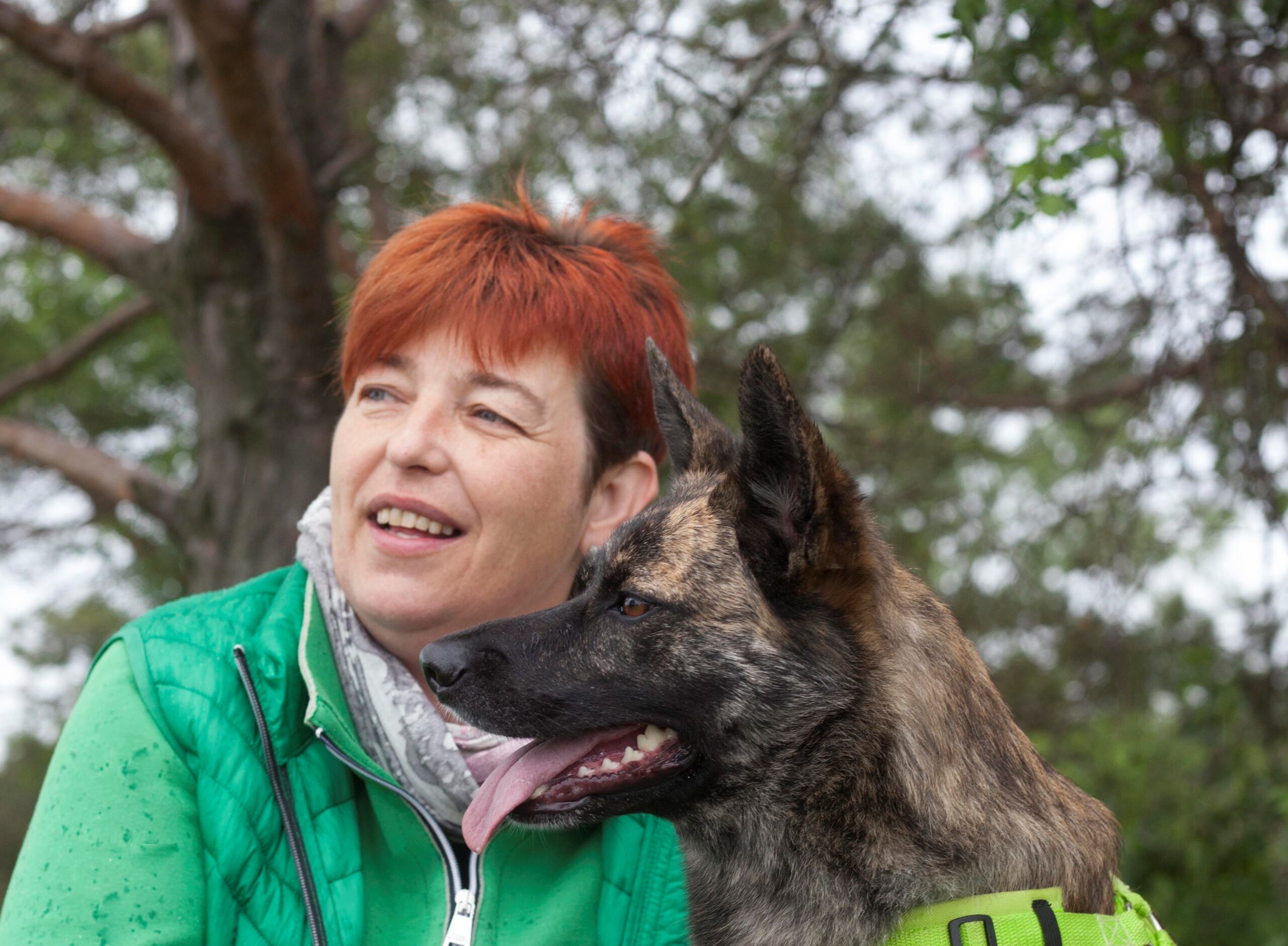 Annette Unterweger in der Natur, blickt lächelnd in die Ferne. Sie hat rotes, kurzes Haar und trägt eine grüne Jacke. Rechts von ihr, ihr Schäferhund Charly.